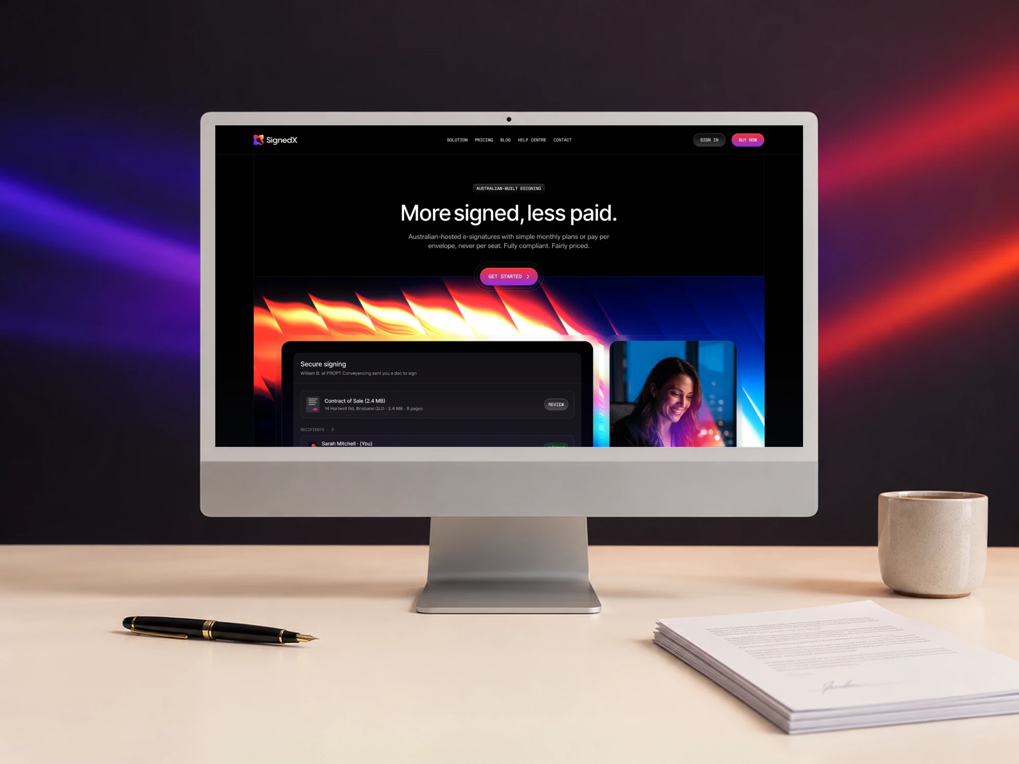 SignedX homepage on a desktop monitor on a light desk with a fountain pen, a ceramic cup and a signed document, against a dark wall lit with purple and red streaks